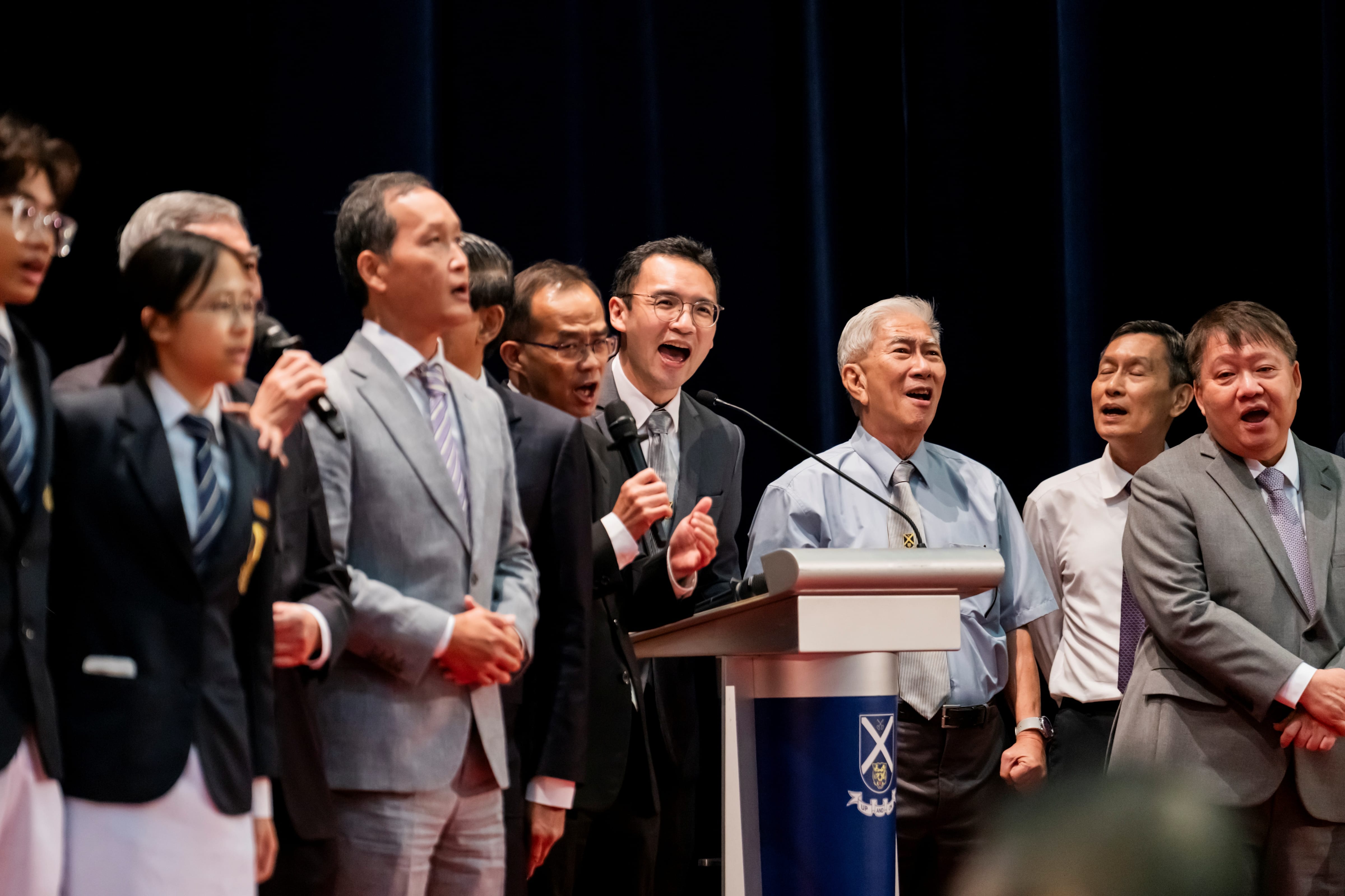 A group of school leaders, guests, and students standing on stage and singing enthusiastically around a podium.